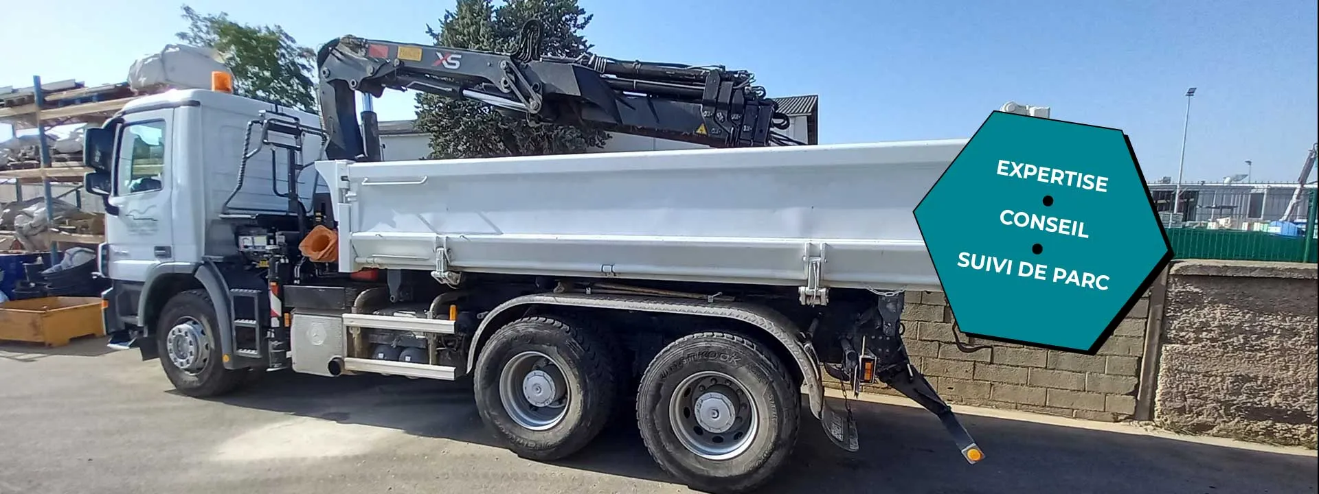 Camion-benne blanc équipé d’une grue auxiliaire stationné en extérieur.